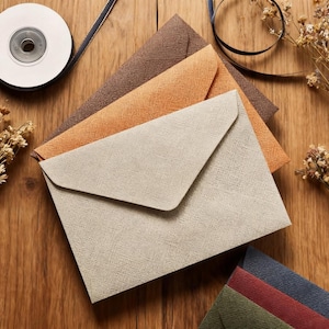 May include: An overhead shot of several textured envelopes in various colors: beige, orange, and brown. A roll of white ribbon and a dark blue ribbon are in the background. Dried flowers are scattered around the envelopes.