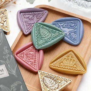 May include: A collection of colorful, triangular wax seals in shades of pink, blue, green, yellow, and purple. Each seal features a unique embossed design, including floral, botanical, and geometric patterns. The seals are arranged on a wooden tray.
