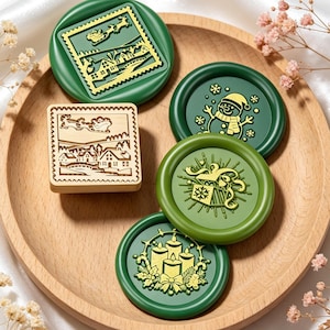 May include: A collection of four circular green wax seals and one square wooden stamp. The seals feature gold-colored embossed designs including a winter village with a sleigh, a snowman, a gift box, and three candles. The wooden stamp shows a matching winter village scene.