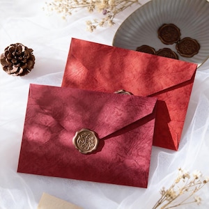 May include: Two red textured envelopes with gold wax seals. The envelopes are sealed with a gold wax stamp that reads "For You" in cursive script. The envelopes are on a white surface.