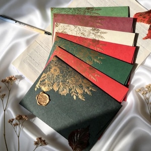 Könnte beinhalten: Eine Sammlung farbenfroher Umschläge mit goldenen floralen Designs und einem Wachssiegel. Die Umschläge sind in Grün-, Rot- und Cremetönen gehalten. Ein Stift und Papier befinden sich im Hintergrund und deuten auf Schreibwaren oder Einladungen hin.