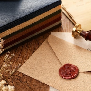 May include: A stack of colorful paper with a textured cover sits next to a tan envelope sealed with a red wax stamp. A brass and wood wax seal stamp and a piece of paper are also visible on the wooden surface. The paper stack has blue, black, green, and brown pages.