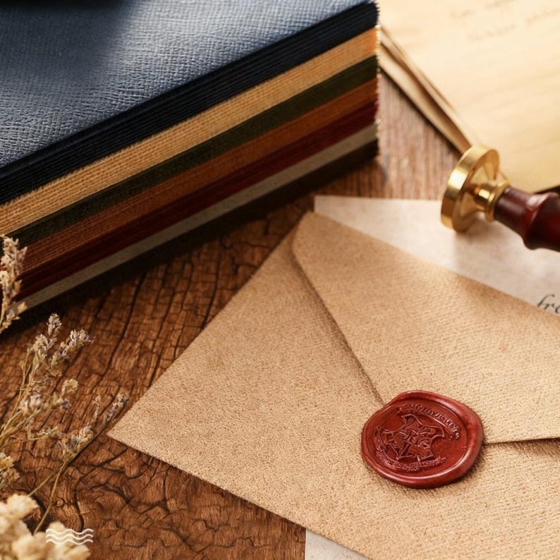 May include: A stack of colorful paper with a textured cover sits next to a tan envelope sealed with a red wax stamp. A brass and wood wax seal stamp and a piece of paper are also visible on the wooden surface. The paper stack has blue, black, green, and brown pages.