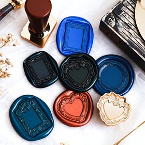 May include: A collection of colorful wax seals in various shapes, including frames and hearts. A wooden stamp with a gold base and brown handle is also present. The seals are in shades of blue, black, red, and gold.