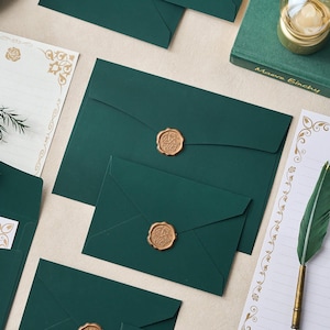 May include: Deep green envelopes sealed with gold wax stamps. The image includes stationery, such as a feather quill and decorative paper with gold detailing. A book titled "Maeve Binchy" is also visible.