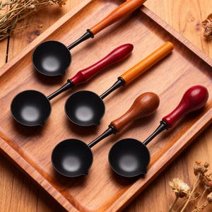 Könnte beinhalten: Eine Sammlung schwarzer Wachsschmelzlöffel mit Holzgriffen in verschiedenen Farben, darunter Natur, Rot und Braun. Die Löffel sind auf einem Holztablett angeordnet und präsentieren ihr Design und ihre Handwerkskunst. Ideal für Bastelarbeiten und Kerzenherstellung.