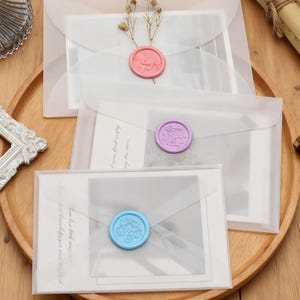 Puede incluir: Tres sobres translúcidos con tarjetas en su interior, sellados con sellos de cera. Los sellos son rosas, morados y azules, cada uno con un diseño de perro. Los sobres están dispuestos sobre una bandeja de madera, con flores secas y elementos decorativos.