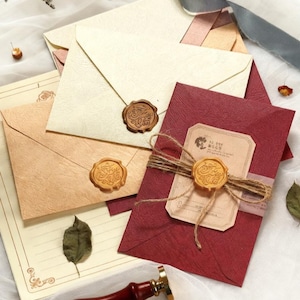 May include: An assortment of stationery items, including cream, tan, and burgundy envelopes, sealed with gold wax stamps. A wooden and gold wax seal stamp is in the foreground. The envelopes are tied with twine and ribbon.