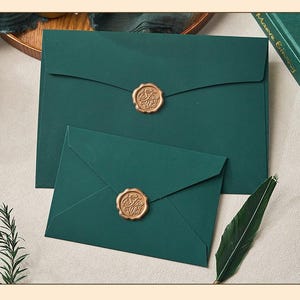 May include: Two dark green envelopes, one large and one small, sealed with gold wax stamps. The stamps feature an ornate design. A green feather and a sprig of greenery are also visible.