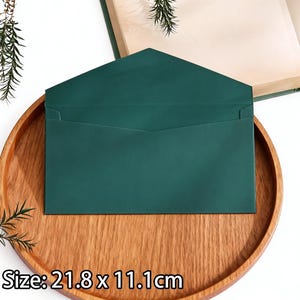 May include: A dark green envelope, measuring 21.8 x 11.1cm, sits on a wooden tray. The envelope has a rectangular shape with a pointed flap. The tray is round and made of light brown wood. The background includes a book and greenery.