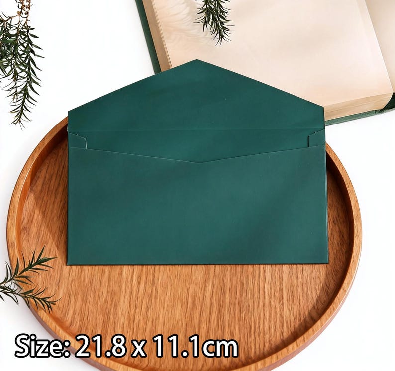 May include: A dark green envelope, measuring 21.8 x 11.1cm, sits on a wooden tray. The envelope has a rectangular shape with a pointed flap. The tray is round and made of light brown wood. The background includes a book and greenery.