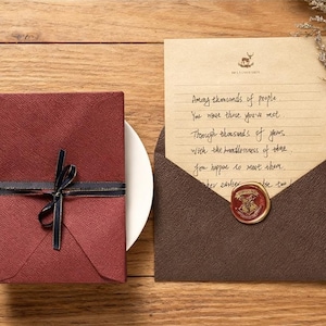 May include: A burgundy gift box tied with a black ribbon and gold trim sits on a white plate. Beside it, an open brown envelope reveals a handwritten letter with a wax seal. The letter's text is partially visible.