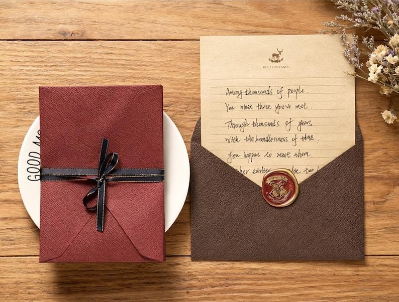 May include: A burgundy gift box tied with a black ribbon and gold trim sits on a white plate. Beside it, an open brown envelope reveals a handwritten letter with a wax seal. The letter's text is partially visible.