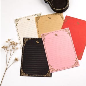 May include: A collection of lined stationery in various colors, including cream, tan, black, pink, and red. Each sheet features a decorative gold border with a bunny illustration. The stationery is arranged on a white surface.