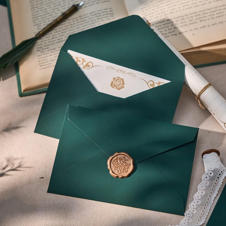 May include: Two dark teal envelopes, one open to reveal a cream card with a gold rose design. The other envelope is sealed with a gold wax seal. A rolled scroll and a book are in the background.