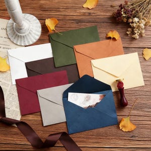 Pode incluir: Uma coleção de envelopes coloridos em várias tonalidades, incluindo branco, castanho, verde e azul, dispostos sobre uma superfície de madeira. Um envelope está aberto, revelando um cartão no interior. Um selo de cera e flores secas também estão presentes.