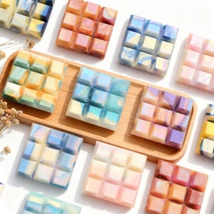 Può includere: Una vista dall'alto mostra diverse cere profumate quadrate e colorate disposte su un vassoio di legno e una superficie bianca. Le cere profumate sono divise in quadrati più piccoli, con colori tra cui blu, rosa, arancione, giallo e viola. Alcune hanno un effetto marmorizzato.