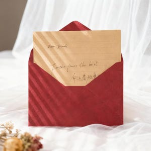 May include: A red envelope with a textured surface, holding a tan-colored card. The card has handwritten text that reads "Dear friends" and other script. The envelope is open, revealing the card inside. The background is a white, sheer fabric.
