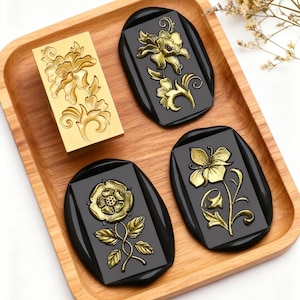 Könnte beinhalten: Ein rechteckiger Wachssiegelstempel aus Messing mit einem komplexen Blumenmuster, begleitet von drei schwarzen Wachssiegeln, die jeweils ein goldenes, geprägtes Blumenmotiv zeigen, alles auf einem hellen Holztablett angeordnet.