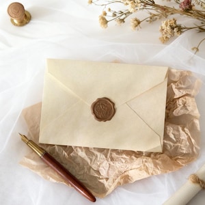 May include: A cream-colored envelope sealed with a brown wax stamp, resting on crumpled brown paper. A wooden and gold fountain pen lies nearby. Dried flowers and a wax seal stamp are also visible, suggesting a vintage or handcrafted theme.