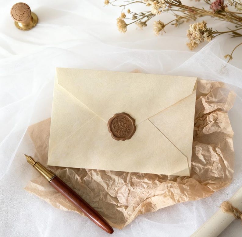 May include: A cream-colored envelope sealed with a brown wax stamp, resting on crumpled brown paper. A wooden and gold fountain pen lies nearby. Dried flowers and a wax seal stamp are also visible, suggesting a vintage or handcrafted theme.