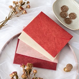 May include: A stack of textured red, pink, and cream-colored envelopes. Dried flowers and a small dish with wax seals are arranged around the envelopes. A gold-colored wax stamp is also visible.