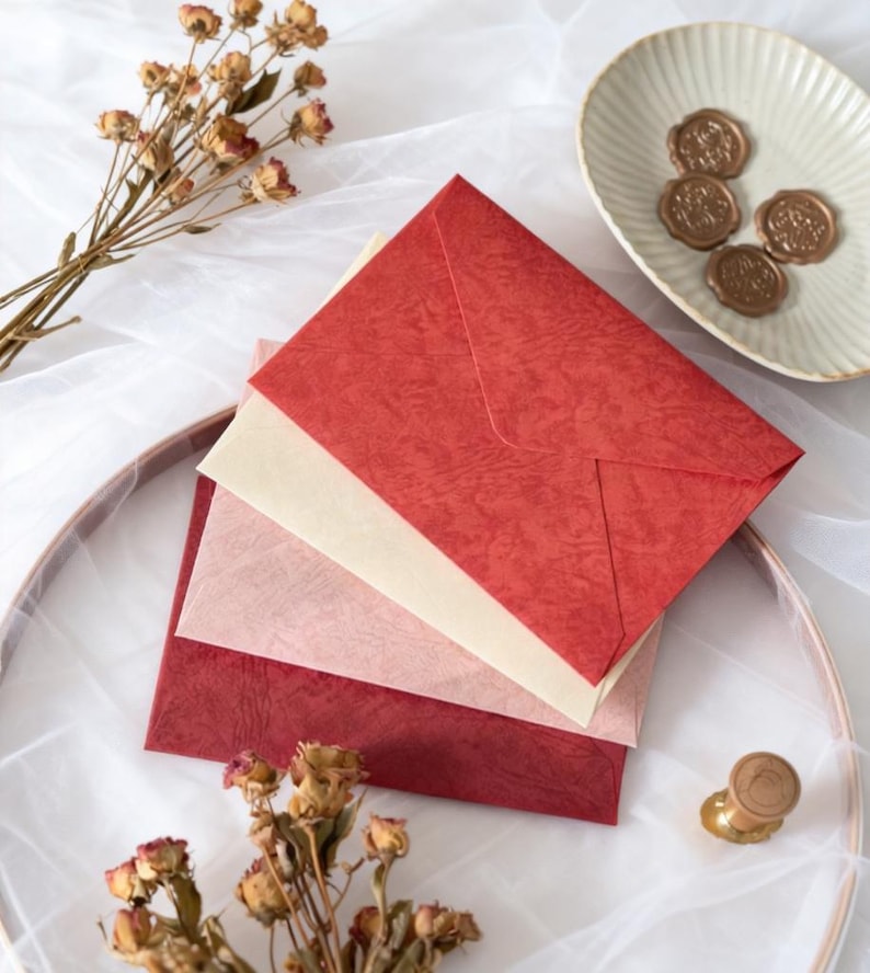 May include: A stack of textured red, pink, and cream-colored envelopes. Dried flowers and a small dish with wax seals are arranged around the envelopes. A gold-colored wax stamp is also visible.