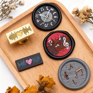 May include: A collection of music-themed wax seals and a stamp, displayed on a wooden tray. The seals feature musical notes, a treble clef, and a heart-shaped sound wave design. Colors include black, red, gold, and gray.