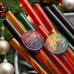 Puede incluir: Una variedad de barras de lacre de colores en rojo, verde, ámbar y negro. Se ven dos sellos de cera decorativos con diseños florales y abstractos. La imagen está ambientada sobre un fondo de adornos navideños y vegetación, sugiriendo un tema festivo.