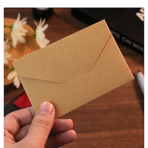 Könnte beinhalten: Ein rechteckiger Umschlag aus hellbraunem Kraftpapier mit einem dreieckigen Verschluss. Der Umschlag hat eine glatte, matte Textur und ein schlichtes, klares Design mit sichtbarer Papierstruktur, dargestellt in einer horizontalen Ausrichtung.