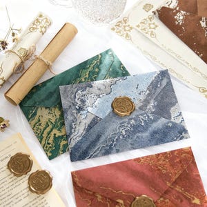 Puede incluir: Surtido de artículos de papelería decorativos, que incluyen sobres veteados en tonos azules, verdes y rojos, sellados con sellos de cera dorados. Un rollo de pergamino enrollado y papel estampado se suman a la estética vintage. La papelería está dispuesta sobre una superficie blanca.