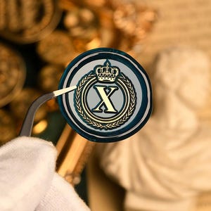 May include: A circular wax seal in gold, featuring a crown and the letter "X" within a laurel wreath. The seal has a dark blue border and is held by tweezers. The design is reminiscent of heraldry.
