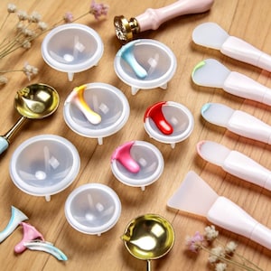 Pode incluir: Uma coleção de materiais de artesanato, incluindo moldes de silicone transparentes, pequenas espátulas e um carimbo de cera. Os moldes contêm cera colorida e as espátulas têm cabos cor-de-rosa. Uma colher dourada e uma colher com cabo rosa também estão presentes.