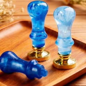 May include: Three blue wax seal stamps with gold-colored bases rest on a wooden tray. The stamps have a marbled appearance, with varying shades of blue. One stamp lies on its side, while the other two stand upright.