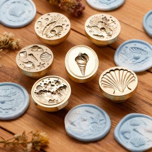 Puede incluir: Una colección de sellos de cera de latón con varios diseños de conchas marinas, incluyendo caracolas y conchas de vieira, junto con sellos de cera azul claro. Los sellos están dispuestos sobre una superficie de madera, con acentos florales secos.