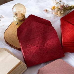 May include: A stack of red textured envelopes with a matching envelope in the foreground. Other envelopes are visible in shades of beige and pink. A small decorative lamp and dried flowers are in the background.