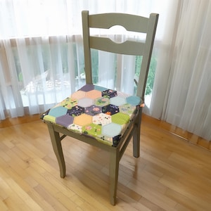 Könnte beinhalten: Ein grüner Holzstuhl mit einem bunten Patchwork-Kissen. Das Kissen besteht aus Stoffquadraten, die in einem sechseckigen Muster zusammengenäht sind. Der Stuhl steht vor einem Fenster mit weißen Vorhängen.