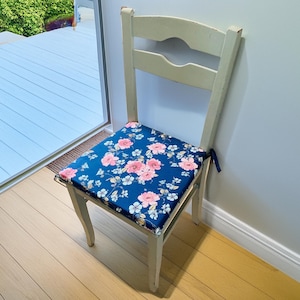 May include: A white wooden chair with a floral patterned cushion. The cushion is blue with pink flowers. The chair is sitting on a wooden floor.