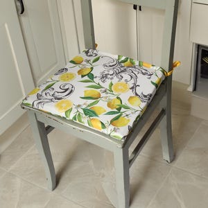 以下が含まれることがあります： レモンと花の模様のクッションが付いた白い椅子。椅子は薄いグレーに塗られており、使い古したような仕上げになっています。