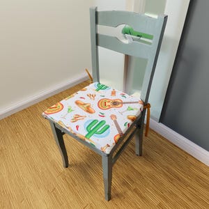 May include: A gray wooden chair with a white cushion featuring a colorful Mexican-themed design. The cushion has images of guitars, sombreros, cacti, and maracas.