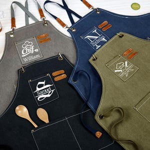 Puede incluir: Varios delantales personalizados en tonos grises, azul marino y verde oliva. Cada delantal tiene un bolsillo y detalles de cuero. Algunos están bordados con nombres y diseños de cocina. Se ven cucharas de madera.