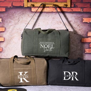 Peut inclure: Trois sacs de sport en toile, vert olive, beige et noir, chacun avec un nom ou des initiales personnalisées en blanc. Le sac vert olive a une bandoulière et le texte "Groomsman Noel Smith". Le sac beige porte le nom "Kevin". Le sac noir porte les initiales "DR Gamble".