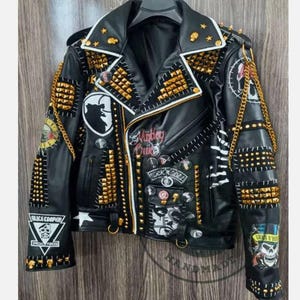 Pode incluir: Jaqueta de moto de couro preto com tachas douradas, patches e correntes. A jaqueta tem fecho de correr e gola branca. Os patches incluem "Police Cooper", "Rock N Roll" e "Chains &  A  Dream".
