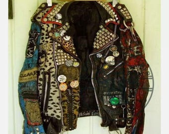 May include: A black leather jacket with a punk rock aesthetic. It is covered in studs, patches, and pins. The jacket has a zip closure and a collar. The patches include a leopard print, a blue and white floral pattern, and a red and white pattern. The jacket has a distressed look.