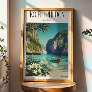 May include: A framed art print of a tropical beach scene with the text "KO PHI PHI DON". The artwork shows a boat on turquoise water, green cliffs, and white flowers. The frame is light brown. The print is displayed on a wooden stool.