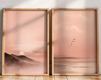 May include: Two framed landscape art prints. The prints depict a sunset over mountains and a body of water with birds in flight. The colour palette is soft, with shades of pink, brown, and white. The frames are made of light-coloured wood.
