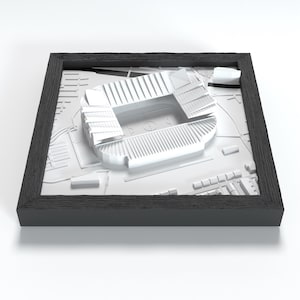 Può includere: Un modello tridimensionale bianco di uno stadio sportivo con gradinate e un campo centrale, inserito in una cornice quadrata in legno nero. Il modello raffigura in dettaglio la struttura architettonica dello stadio e dell'area circostante.