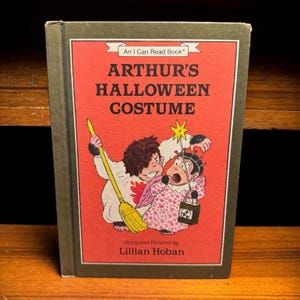 May include: A hardcover book titled Arthur's Halloween Costume by Lillian Hoban. The red cover features an illustration of two characters in costumes, one holding a yellow broom and the other a star wand and a small black bucket.