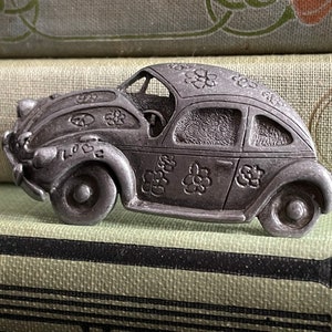 May include: A pewter figurine of a Volkswagen Beetle car with a peace sign and flowers on the side. The car is painted in a silver color and has a detailed design.