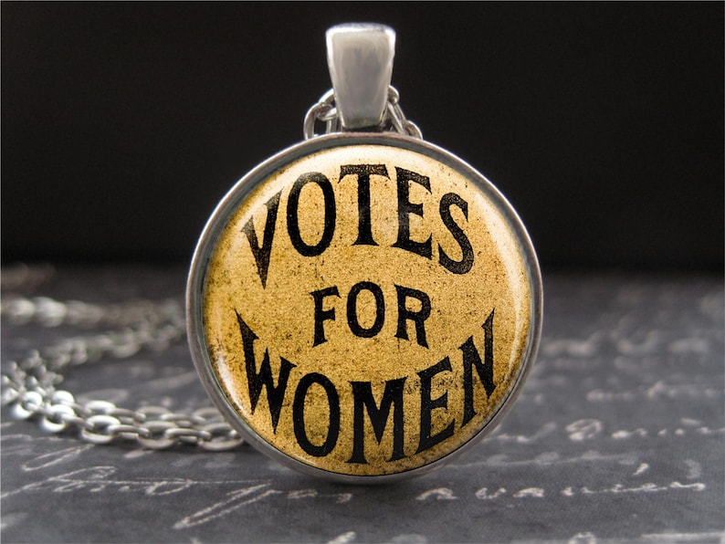 以下が含まれることがあります： 黄色い背景に黒い文字で「Votes for Women」と書かれた丸いペンダントが付いた銀色のネックレスです。